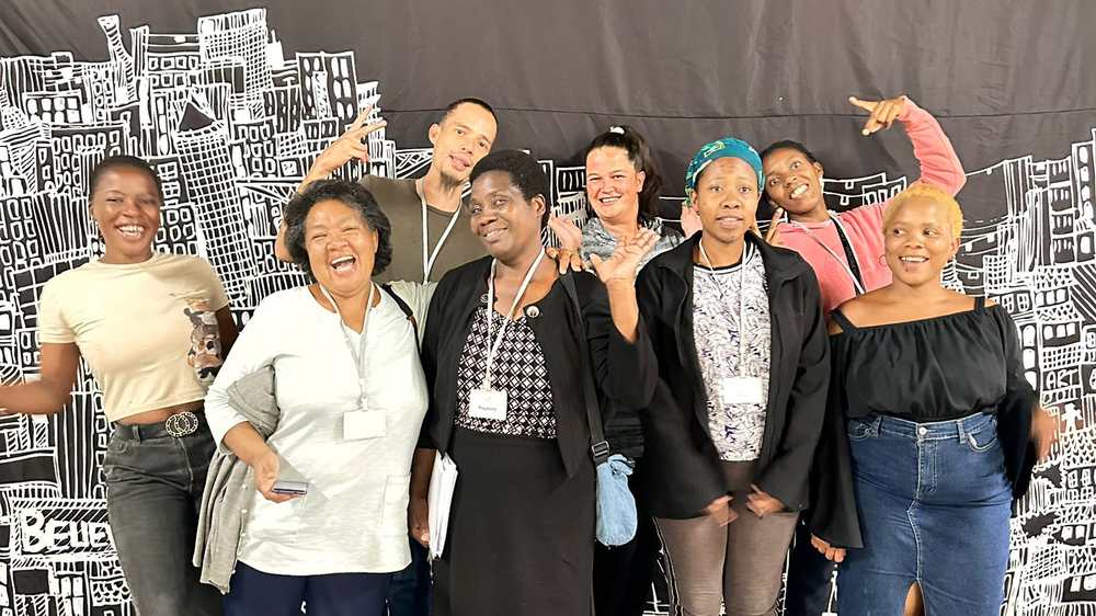 Eight Izenzo Job Club members standing together laughing and posing in front of a large black-and-white city mural, wearing name badges.
